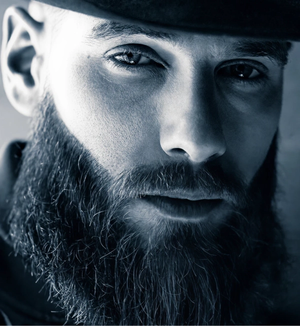 Man with a full beard wearing a stylish hat, demonstrating the use of premium beard care sets and brushes designed for grooming and styling.